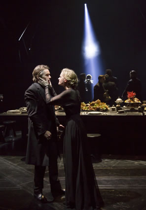 Lady Macbeth has her hands on Macbeth's cheeks, in the background is the banquet table and lords are milling in the darkness with a single shaft of light shining down